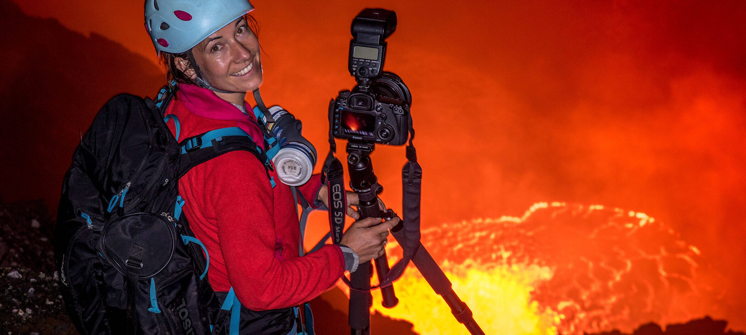 Expediční fotografka Ulla Lohmann stojí s helmou a batohem na okraji sopky a usmívá se na přihlížející. Vedle ní stojí fotoaparát na stativu.