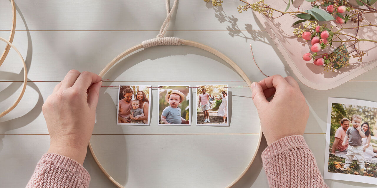 Obrázek dvou rukou upevňujících hotové fotografie na nit v dřevěném kruhu. V pozadí jsou další dřevěné kruhy, květiny a jedna fotka.