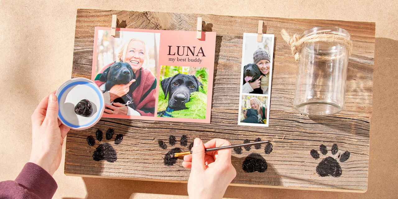 Dvě ruce malují malé psí tlapky na dřevěnou desku černou barvou.
